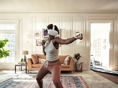 A imagem mostra uma mulher em um sala com um sofá ao fundo brincando com um jogo de realidade virtual. Ela está com um óculos para jogar e os controles em suas mãos