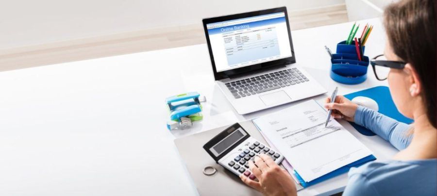 Imagem de uma mulher com a mão esquerda em uma calculadora e com uma caneta na mão direita. a frente a um notebook e diversos itens de escritório. 